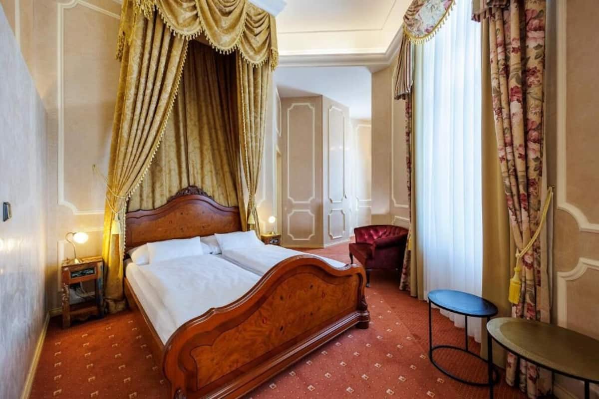 A hotel room with a wooden bed, a small bedside table, a chair, and a large window with curtains.