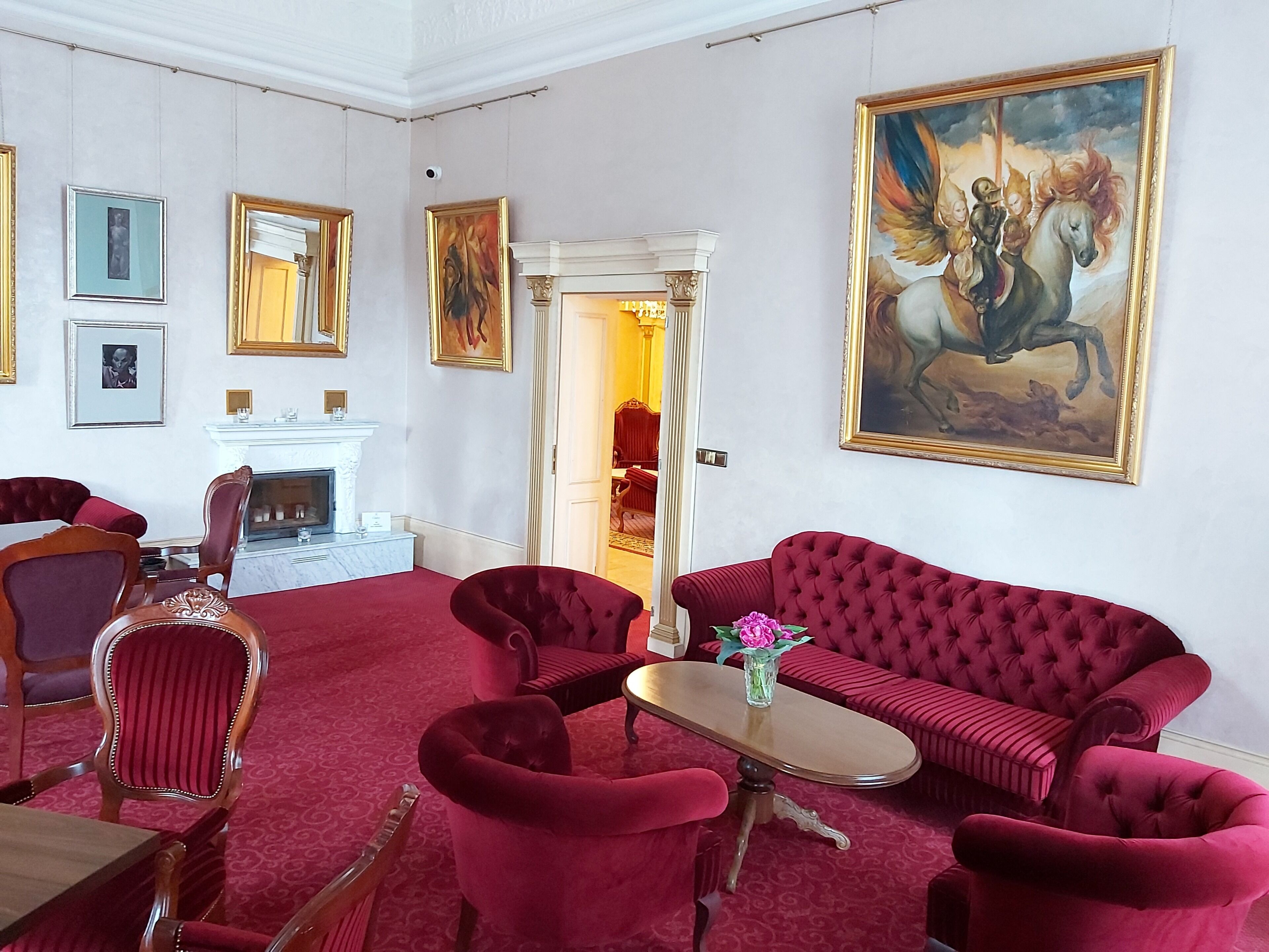 A room with a red sofa, matching chairs, a small table, and a fireplace. There are framed pictures on the walls and a large painting of a horse.