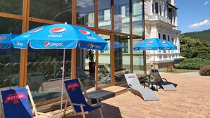 Un patio de hotel con mobiliario exterior, incluyendo sillones reclinables y sombrillas, y un edificio de vidrio grande al fondo.