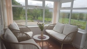 A sunlit room with wicker furniture, a small table, and a view of a green landscape through large windows. - Y Big (Pwllheli)
