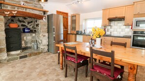 A kitchen with a stone wall, wooden cabinets, a refrigerator, a microwave, and a dining table with chairs. - Y Big (Pwllheli)