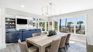 A modern living room with a blue sofa, a wooden dining table with chairs, and a large window offering a view of the beach.