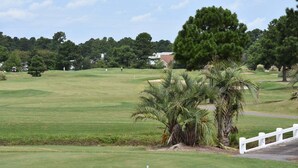 A golf course with a white fence, palm trees, and a building in the background. - Close to Beach Brunswick Plantation Condo 2302M with 27 Hole Golf Course Onsite by RedAwning (Calabash)