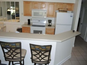 A kitchen with wooden cabinets, a microwave, a stove, and a refrigerator. There are two chairs with a decorative backrest.