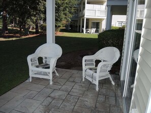 A porch with two wicker chairs and a small table, a grassy yard, and a building in the background.