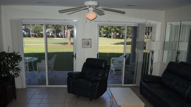 A living room with a ceiling fan, a black leather sofa, a black armchair, and a glass door leading to a patio with outdoor furniture. - Close to Beach Brunswick Plantation Condo 2302M with 27 Hole Golf Course Onsite by RedAwning (Calabash)