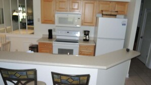A kitchen with wooden cabinets, a microwave, a stove, and a refrigerator. There are two chairs with a decorative backrest. - 2302 2 Bedroom Villa by RedAwning (Calabash)