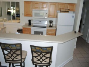 A kitchen with wooden cabinets, a microwave, a stove, and a refrigerator. There are two chairs with a decorative backrest. - 2302 2 Bedroom Villa by RedAwning (Calabash)