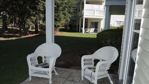 A porch with two wicker chairs and a small table, a grassy yard, and a building in the background. - 2302 2 Bedroom Villa by RedAwning (Calabash)