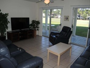 A living room with a television, a sofa, a coffee table, and a chair. There is a plant and a view of the outdoors through a glass door. - 2302 2 Bedroom Villa by RedAwning (Calabash)