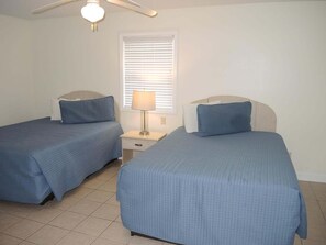 A bedroom with two beds, a ceiling fan, a window with blinds, and a bedside lamp.