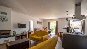 Amplia sala de estar con un sofá amarillo, chimenea, una mesa de comedor y una zona de cocina con fregadero y armarios.
