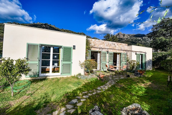 A two-story building with a stone facade, green shutters, and a stone pathway leading to a seating area.