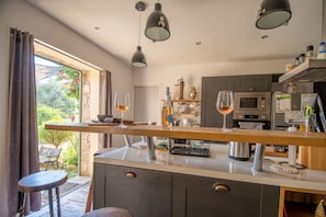 A modern kitchen with a central island, overhead pendant lights, and a view of the garden through the window.