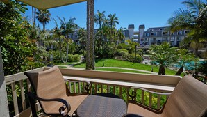 A balcony with wicker chairs and a table, overlooking a well-maintained lawn and a pool area, with a multi-story building in the background. - Tranquil NCV Beach Condo with Lush Gardens & Ocean Glimpses! (Oceanside)