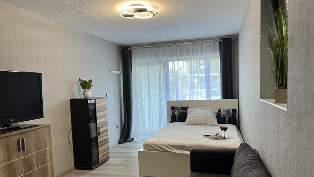 A modern hotel room with a flat-screen TV, a wooden wardrobe, a bed with a white duvet, a dark sofa, and a large window with sheer curtains. - Quiet 1 room apartment (Weilrod)