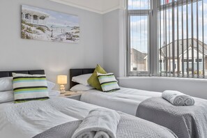 A hotel room with two beds, a painting of a beach scene, a window with blinds, and a view of houses outside. - Plymouth-fernhurst- 2 Bedroom Bungalow (Plymouth)