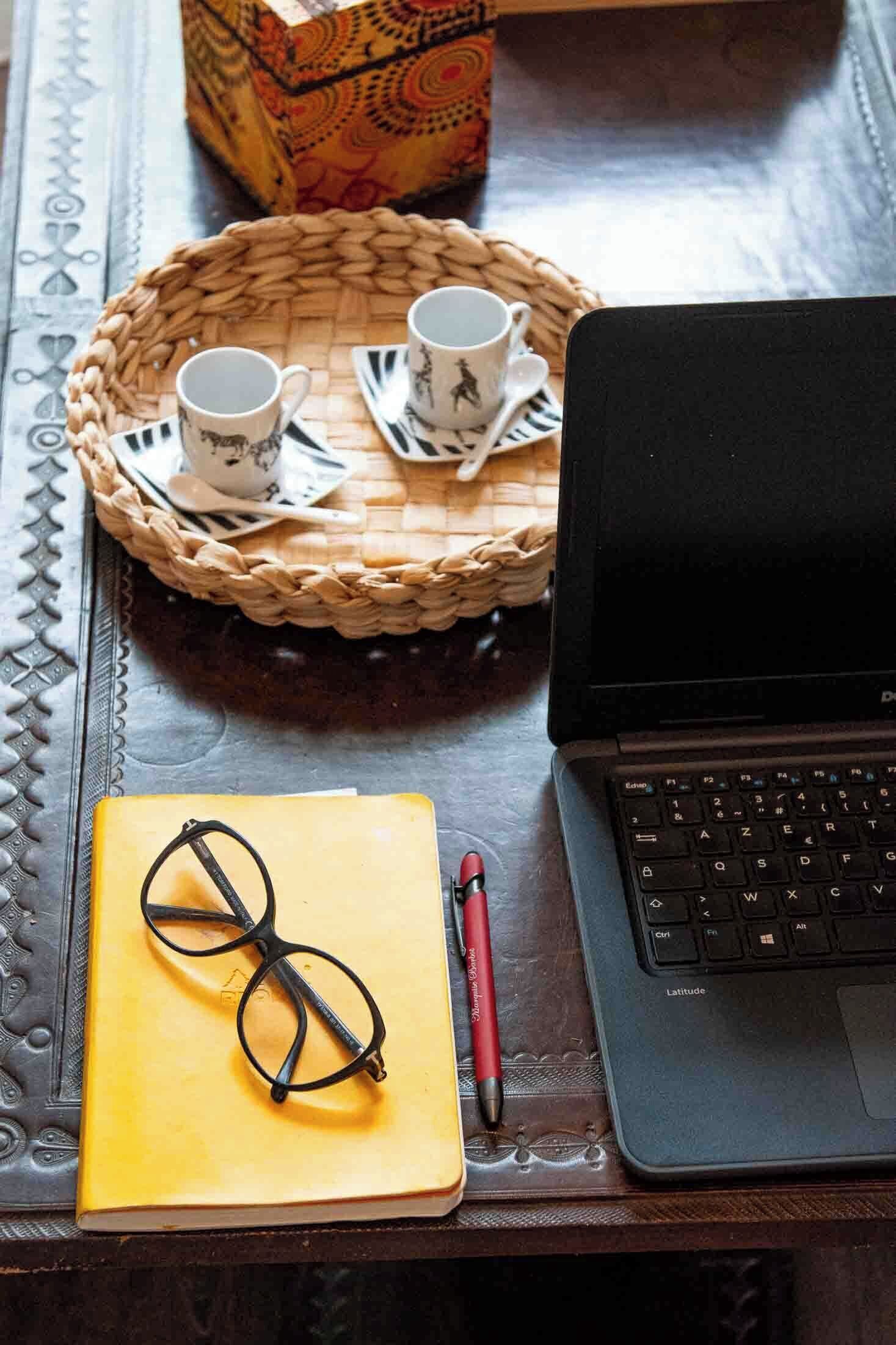 A laptop, a yellow notebook, glasses, a red pen, and a woven tray with cups and saucers on a wooden table.