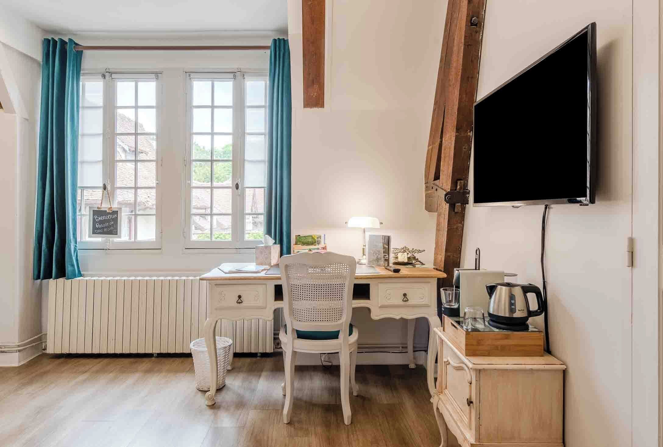Une pièce confortable avec un bureau blanc, une chaise, un radiateur, un téléviseur et une fenêtre avec des rideaux.