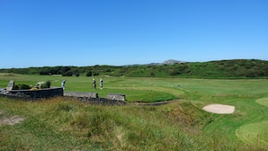A golf course with a stone wall, a flag, and a distant mountain. - Swn Y Nant (Holyhead)