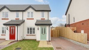 A two-story residential building with a red and green front door, a brick pathway, and a wooden fence. - Swn Y Nant (Holyhead)