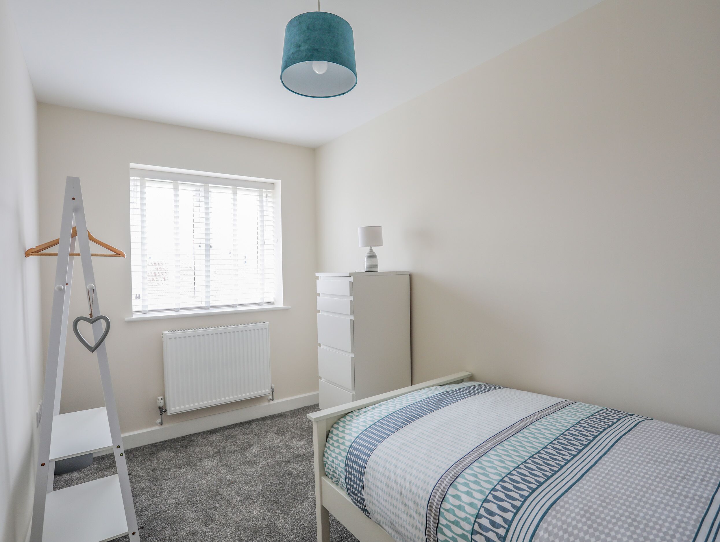 A bedroom with a bed, a dresser, a radiator, a window with blinds, a hanging heart-shaped decoration, and a white ladder.