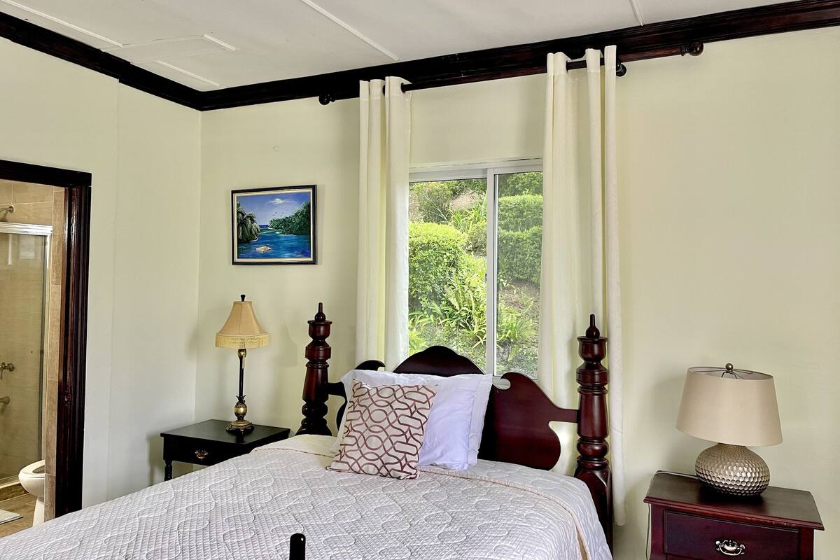 Un dormitorio con cama, mesita de noche, lámpara, ventana con cortinas y un cuadro en la pared.