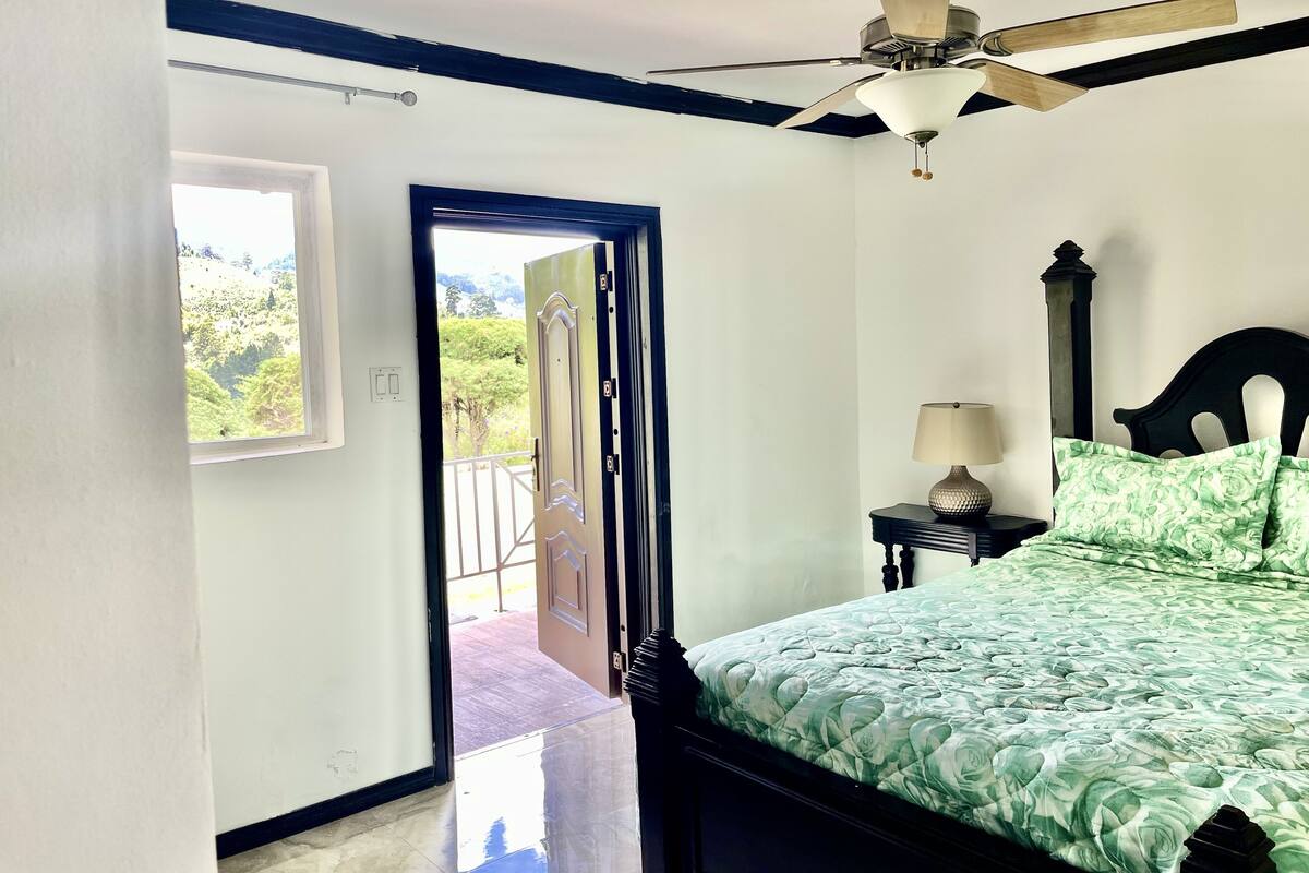 Un dormitorio con cama, mesita de noche, lámpara, ventilador de techo y vistas a un balcón exterior por una puerta abierta.