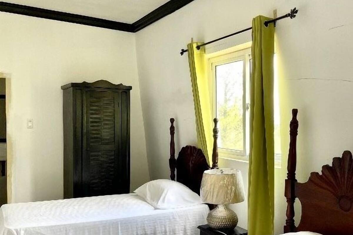 Una habitación con dos camas individuales, un armario de madera, una mesita de noche con lámpara y una ventana con cortinas amarillas.
