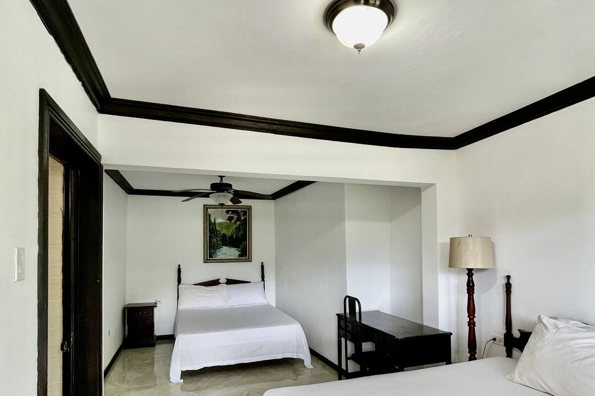Habitación de hotel con una cama grande, una cama más pequeña, un escritorio, una silla, una lámpara y un ventilador de techo.