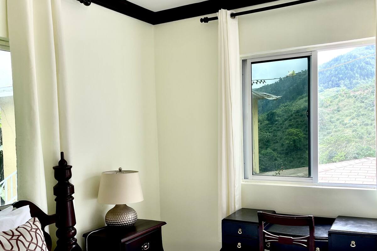 Un dormitorio con cama, escritorio, silla, lámpara y ventana con vistas a un bosque.