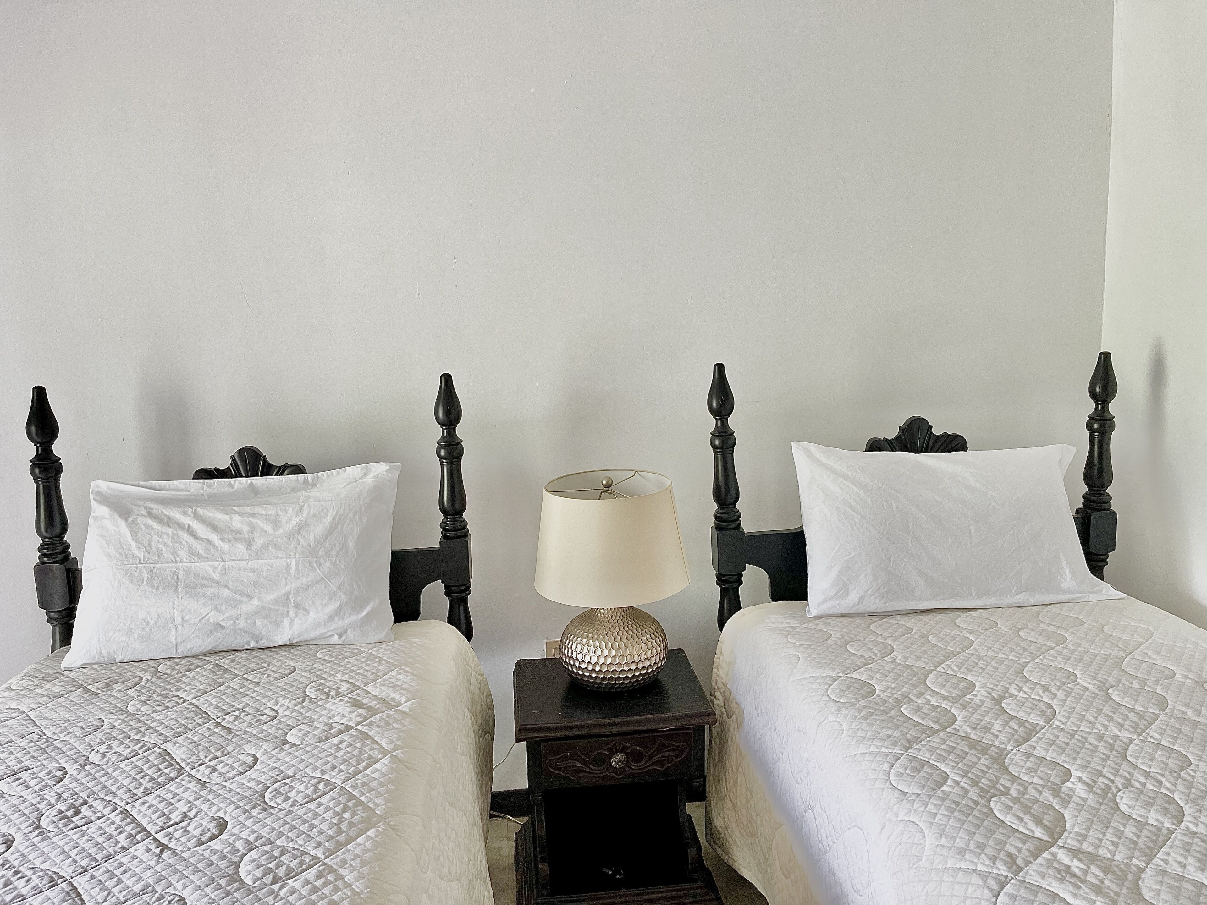 Una habitación con dos camas, cada una con una colcha blanca acolchada y almohadas blancas, flanqueadas por cabeceros de madera oscura. Una mesita de noche con una lámpara y un adorno se encuentra entre las camas.