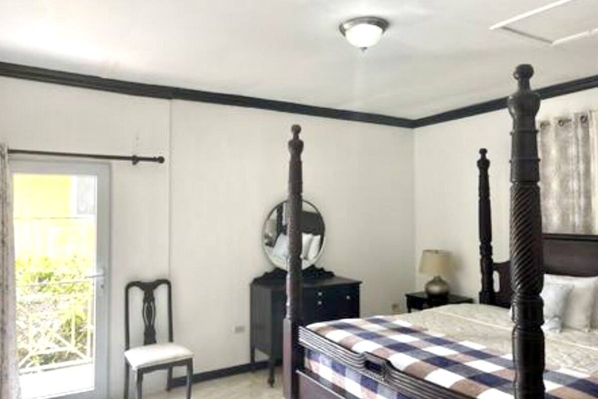 Un dormitorio con cama con dosel, una mesita de noche con lámpara, una silla y una ventana con vistas a zonas verdes.