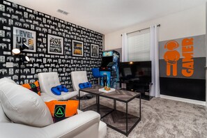 Uma sala de estar com tema de jogos, decorada com ícones de consoles e equipada com uma poltrona gamer, uma mesa de centro e uma grande TV.