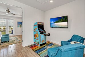 A room with a retro arcade machine, a blue sofa, a colorful rug, and a beach-themed painting. - Last Minute Disney Getaway! Villa with Private Pool & Hot Tub. Book Now! #39456 (Kissimmee)