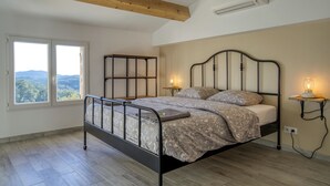 A bedroom with a metal bed frame, a wooden ceiling beam, an air conditioner, a bedside table with a lamp, and a window with a view of the outdoors. - Cosy ALL NEW cottage with swimming pool- (Tanneron)