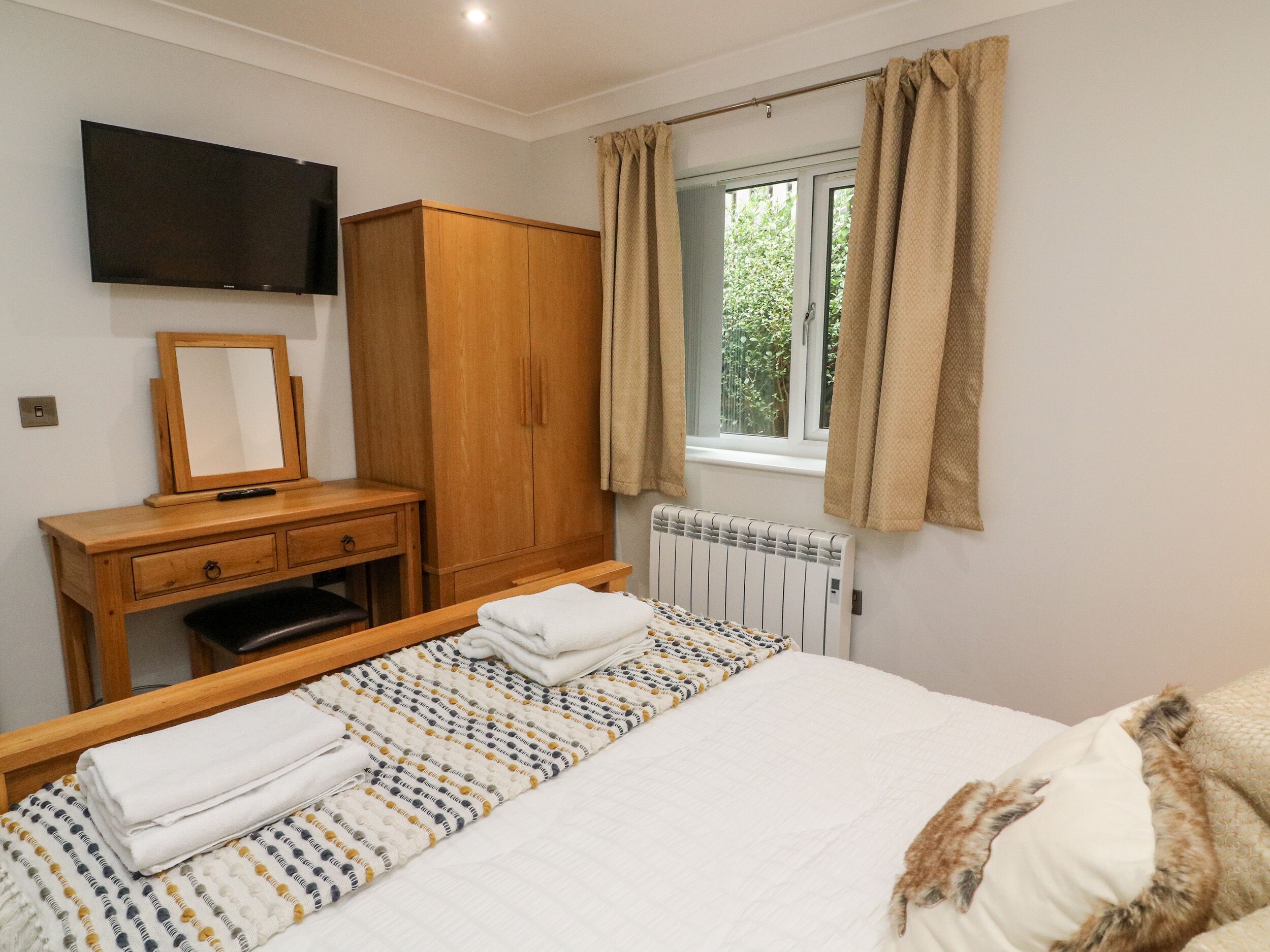 A bedroom with a bed, a wooden wardrobe, a mirror, a radiator, and a window with curtains.