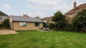 Un jardin arrière d’une maison, avec une pelouse bien entretenue, un espace patio aménagé avec des sièges, et une maison dotée d’un toit en tuiles et de murs en briques.