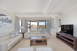 A spacious living room with a large window, a flat-screen TV, a wooden coffee table, and a beige sofa. - Island View 35 (Cape Town)