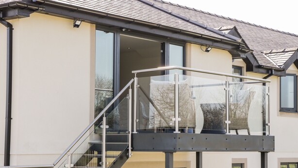 Une maison moderne à deux étages, dotée d’un balcon en verre, d’un escalier apparent et d’une grande fenêtre.