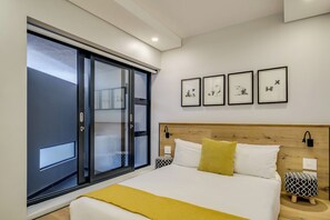 Una habitación de hotel moderna con un gran ventanal corredero, cabecera de madera, cama con ropa de cama blanca y amarilla, y cuatro obras de arte enmarcadas en la pared.