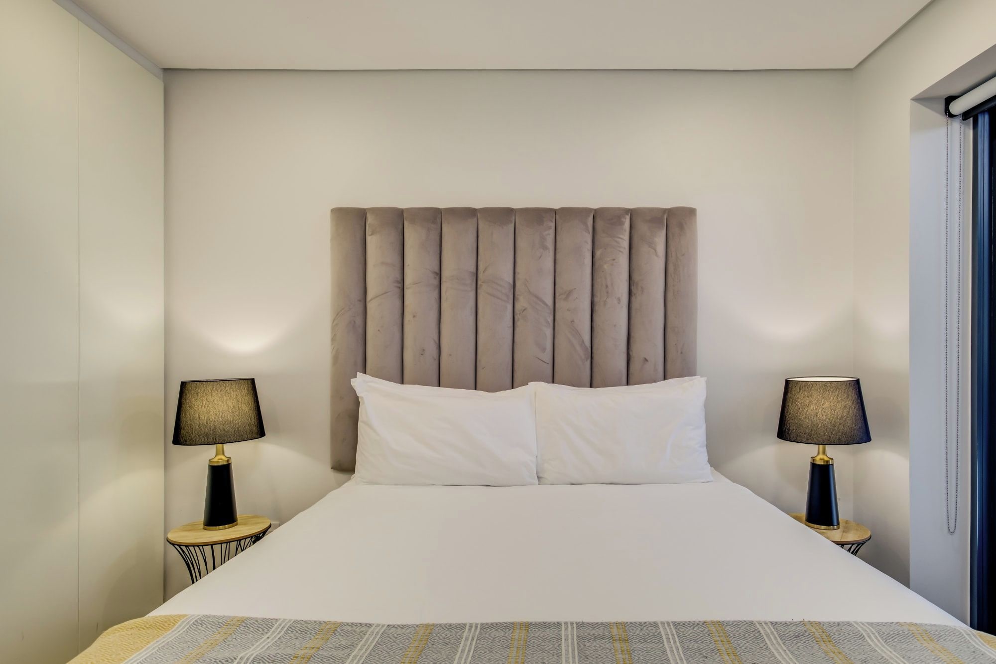 A hotel room with a large bed, two bedside tables with lamps, a headboard, and a window with curtains.