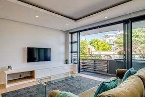 A modern living room with a flat-screen TV mounted on a white console, a glass coffee table, and a sofa with cushions. - Conveniently Located Apartment in Green Point (C on S) (Cape Town)
