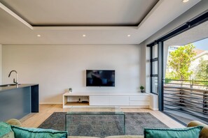 A modern living room with a flat-screen TV mounted on the wall, a built-in kitchen counter, and a large window offering a view of the outdoors. - Conveniently Located Apartment in Green Point (C on S) (Cape Town)