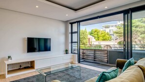 A modern living room with a flat-screen TV mounted on a white console, a glass coffee table, and a sofa with cushions. - Conveniently Located Apartment in Green Point (C on S) (Cape Town)