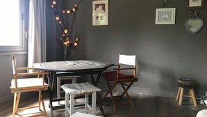 A room with a table, two chairs, a stool, and a small stool with a heart-shaped frame. There are string lights on a branch, and several framed pictures on the wall.