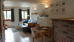 Una sala de estar con sofá, mesa de centro y comedor con mesa y sillas. Hay ventanas con cortinas, una chimenea y cuadros en la pared.