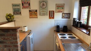 Een compacte keuken met een bakoven, een spoelbak en een fornuis. Er hangen diverse decoratieve borden aan de muur, en er is een raam met zonwering.
