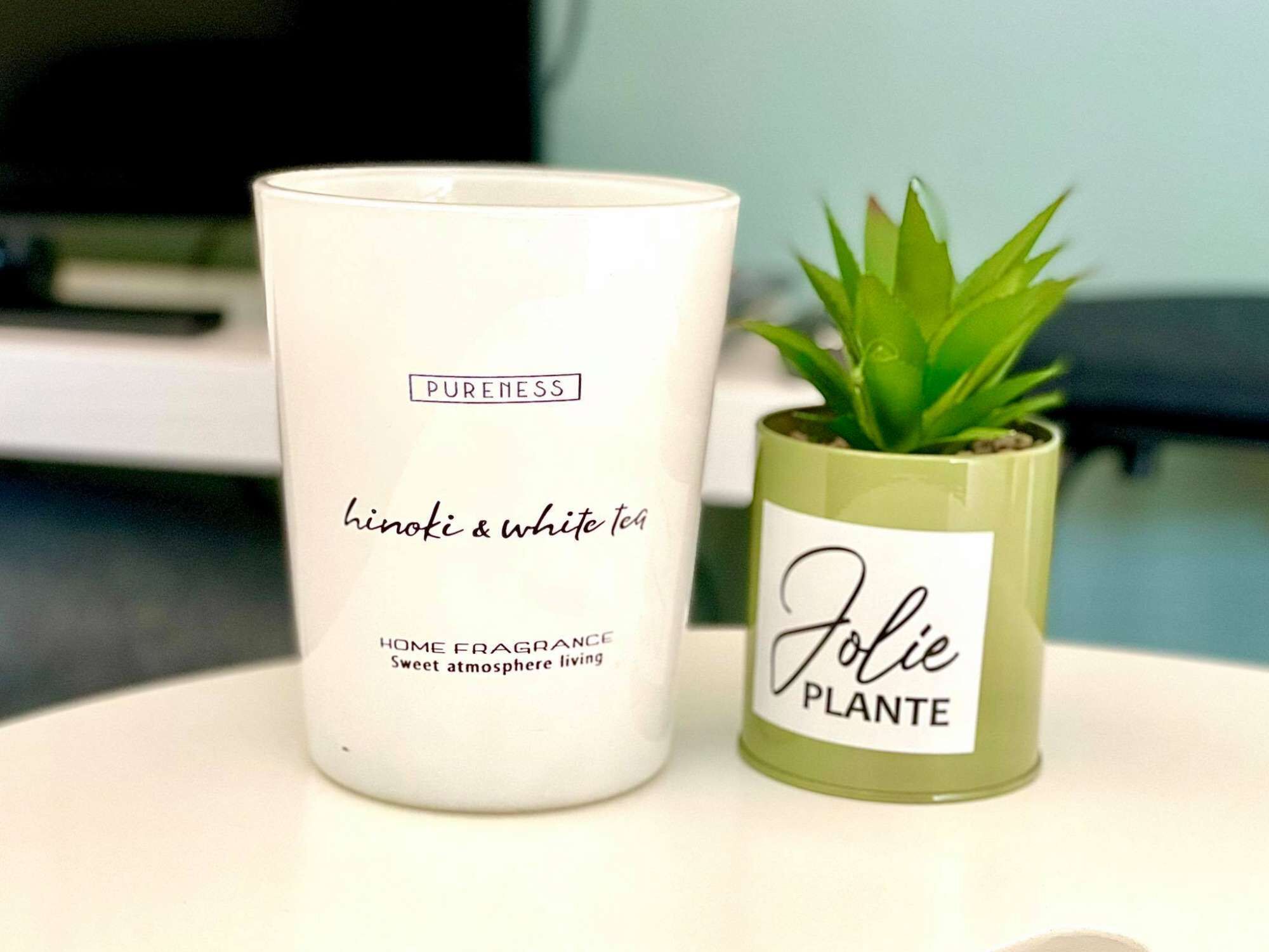 Et hvidt stearinlys med teksten 'PURENESS' og 'hinoki & white tea', samt en lille grøn plante med mærkatet 'Jolie PLANTE' på en hvid overflade.