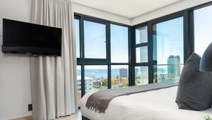 A modern bedroom with a large bed, a flat-screen TV mounted on the wall, and a view of the city through large windows. - One Calais | Corner Studio with Ocean Views (Cape Town)